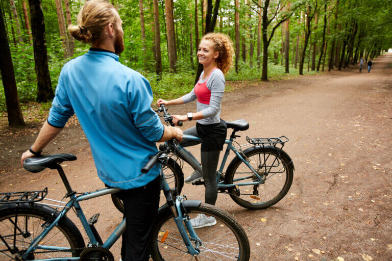 Happy young active couple with bicycles having talk while standing on forest road on summer deay