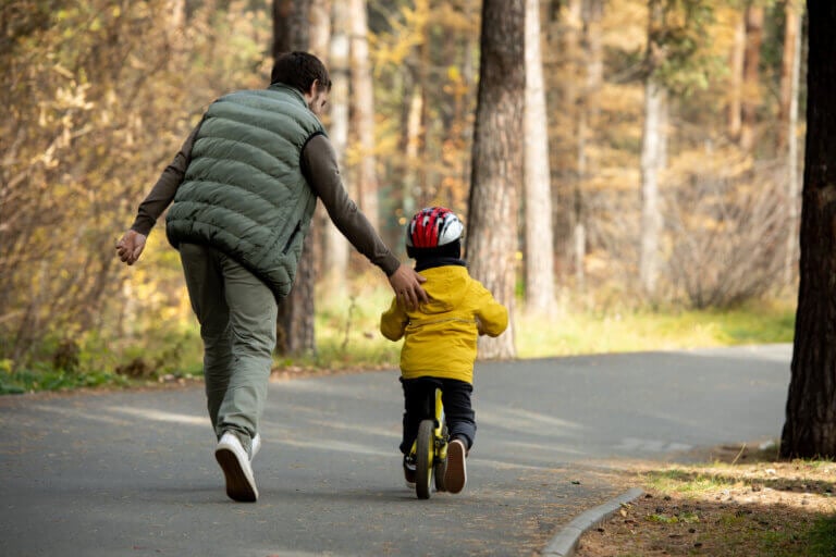 Rear view of young father running after his little son in safety helmet and casualwear while teaching him how to ride balance bicycle in park